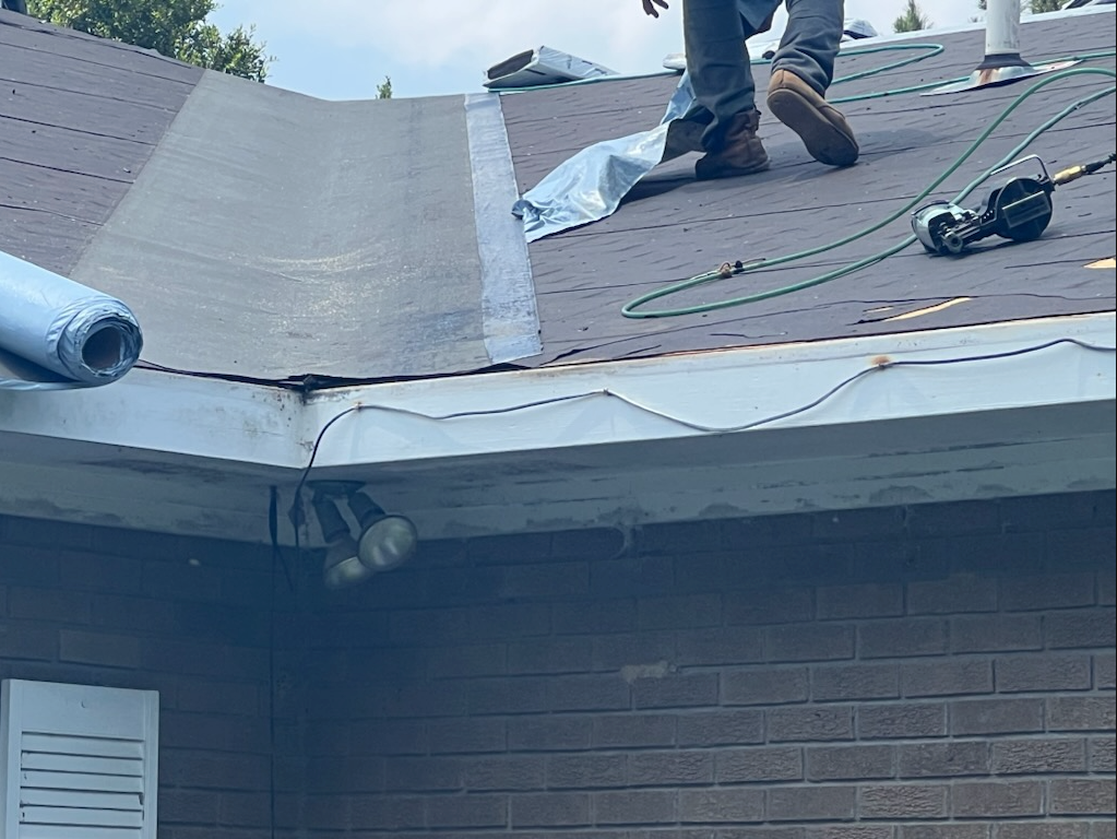 Roofers working on a shingled house roof, with tools and rolled roofing visible.