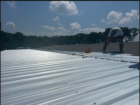 White metal roof under a blue sky with clouds, with a curved vent and treeline in the distance