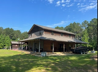 Rustic two-story house with a large wraparound porch, parked truck, and trees under a blue sky