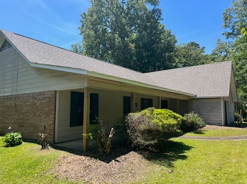 Single-story brick house with a covered porch, gray roof, and trimmed bushes in a sunny yard