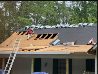 Roof under repair with worker on plywood decking and ladder leaning against the house