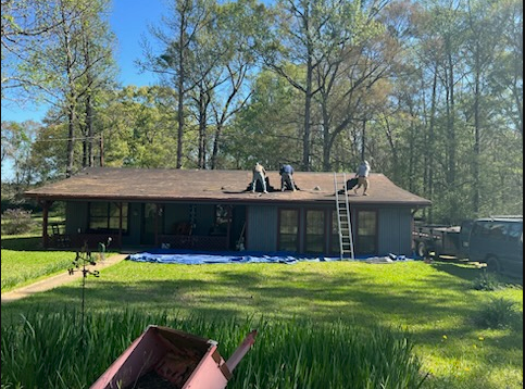 Workers on a roof with a ladder, beside a single-story house surrounded by trees and lawn.