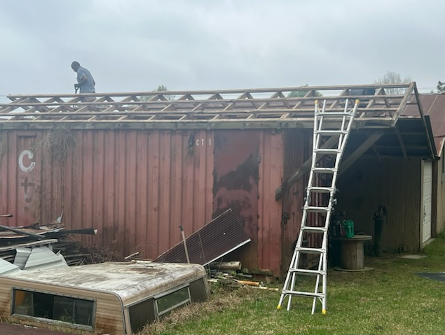Person on a roof beside a ladder, working on a weathered red shed with a partially covered opening.