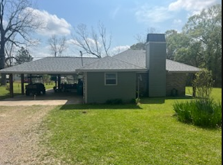 Single-story gray house with carport, brick chimney, and green lawn under a sunny sky
