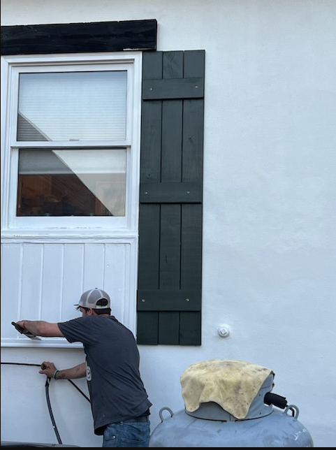 Worker painting a white house exterior beside a window and dark shutters