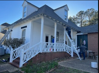White house porch with crisscross railings; a person stands near the front steps on a sunny day.