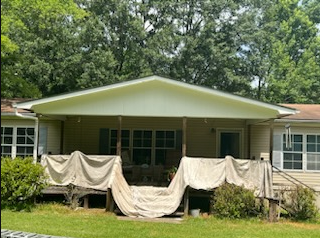 Single-story house with covered porch and draped fabric, framed by trees and grass.