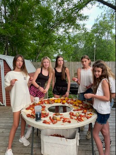 Five people standing around a table covered with colorful food outdoors under trees