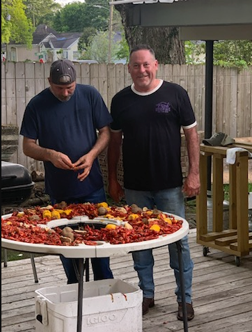 Two men standing by a large seafood boil on a backyard deck, one looking down at the food.