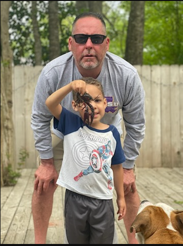 Man standing behind child and dog on a wooden deck, both facing the camera, outdoors near a fence.