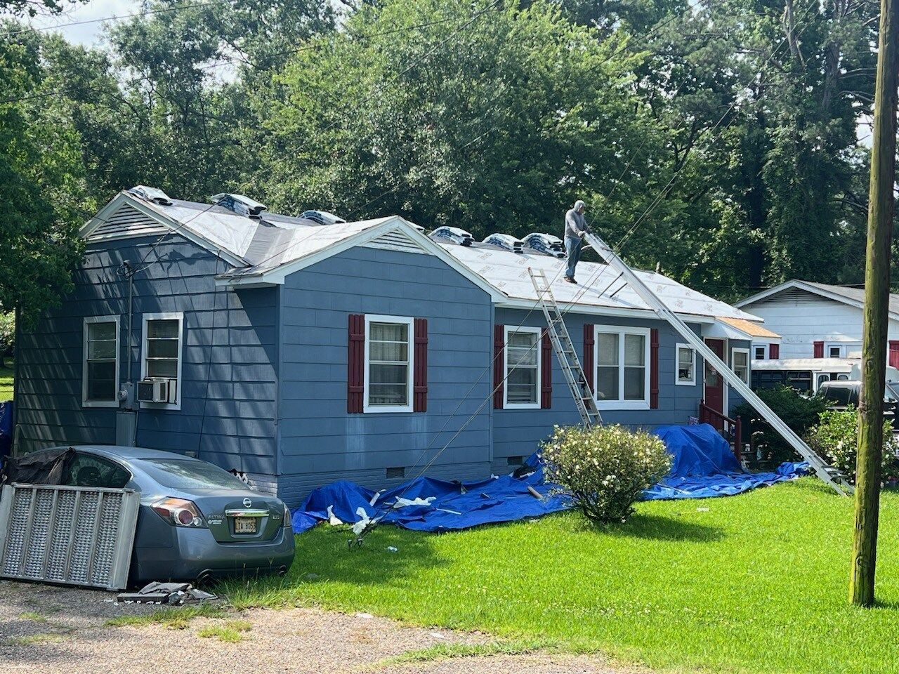 Blue house with storm damage, torn roof, and blue tarp in a grassy yard with trees behind it