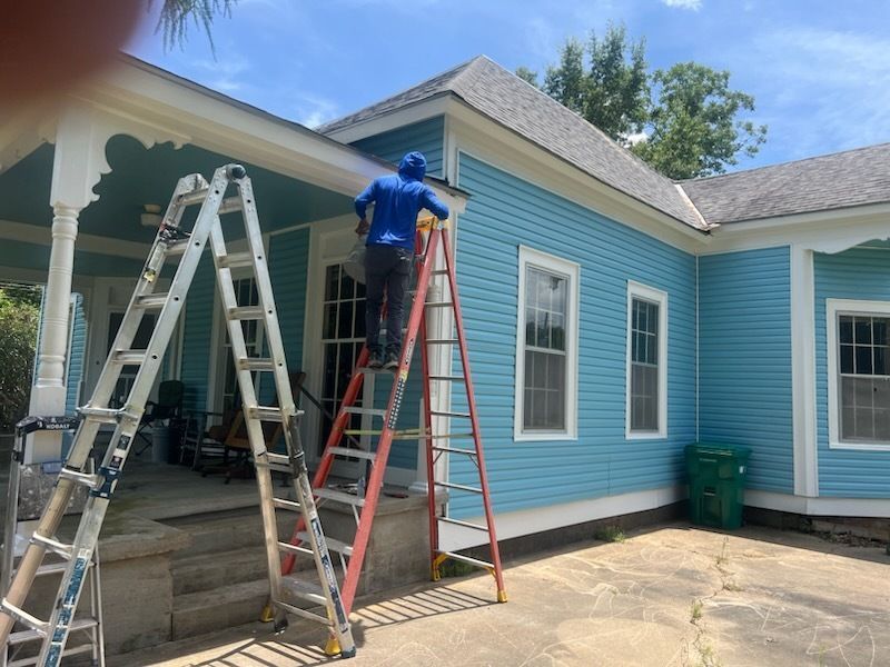 Workers on ladders painting a blue house exterior under the porch roof.