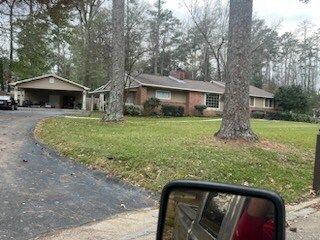 Suburban brick house with a curved driveway, large trees, and a car side mirror in the foreground