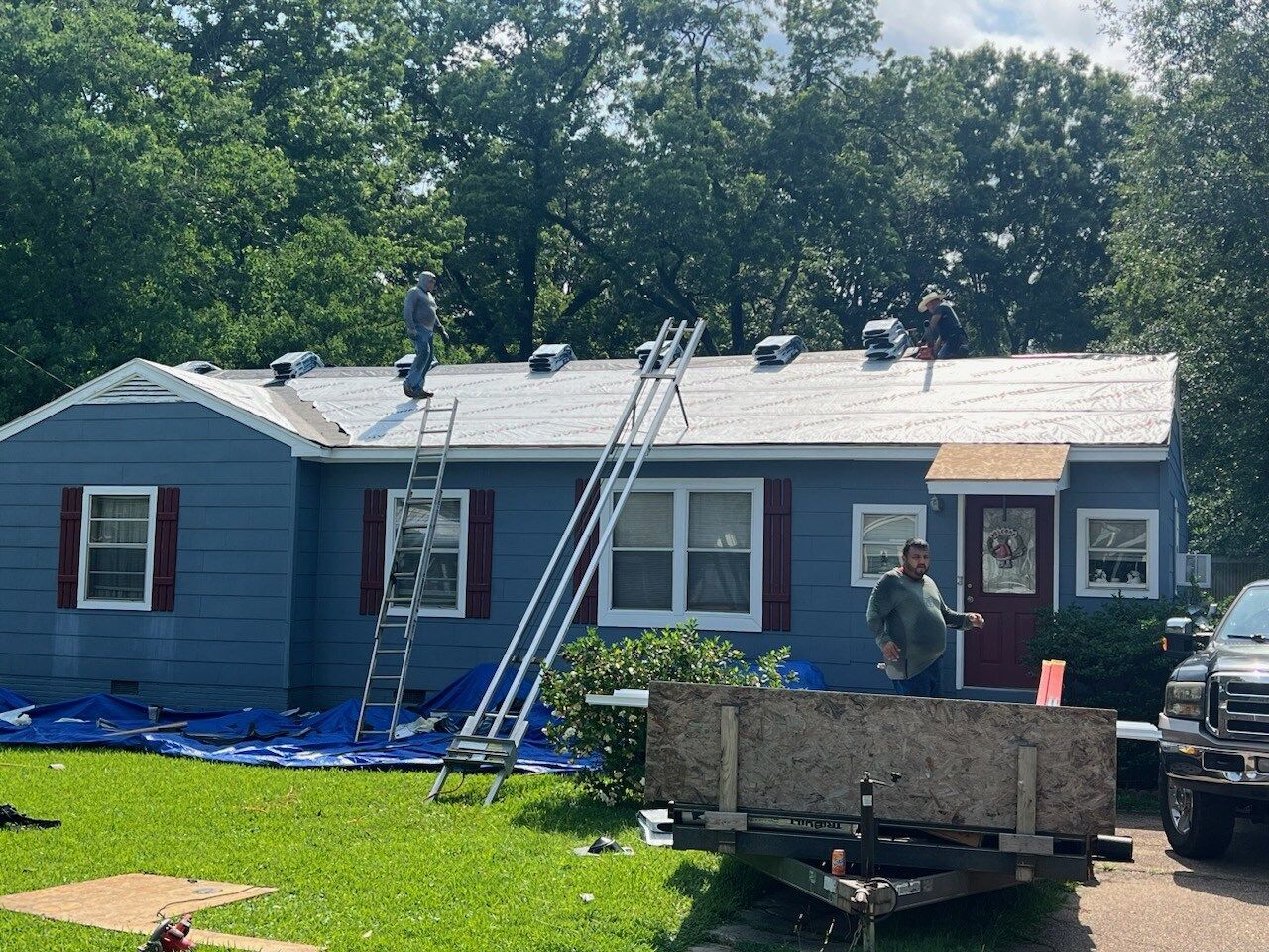 Workers repairing the roof of a blue house, with ladders and tools in the yard.