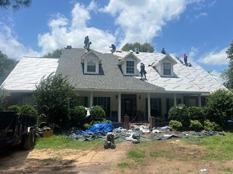 House with white roof under repair, workers on the roof, blue sky and yard debris in front