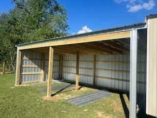 Open-sided carport with wooden posts, corrugated metal roof, and white walls beside grass and trees.