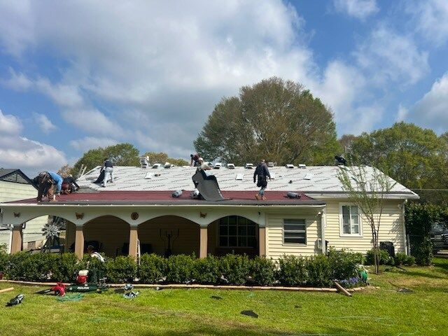 Workers on a house roof, replacing white shingles on a single-story home with a porch.