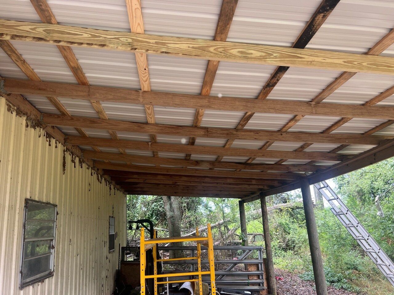 Covered porch with wooden beams, corrugated roof, and yellow scaffolding beside a metal-sided wall outdoors