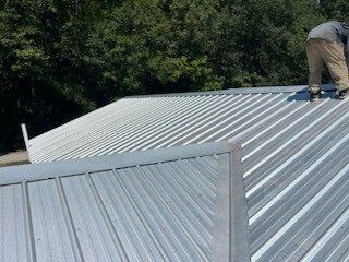 Worker on a corrugated metal roof, installing panels near a tree line.