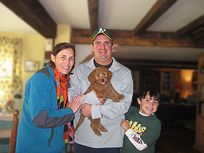 Family with a brown puppy inside a house. Woman in blue, man holding puppy, child smiling in green.