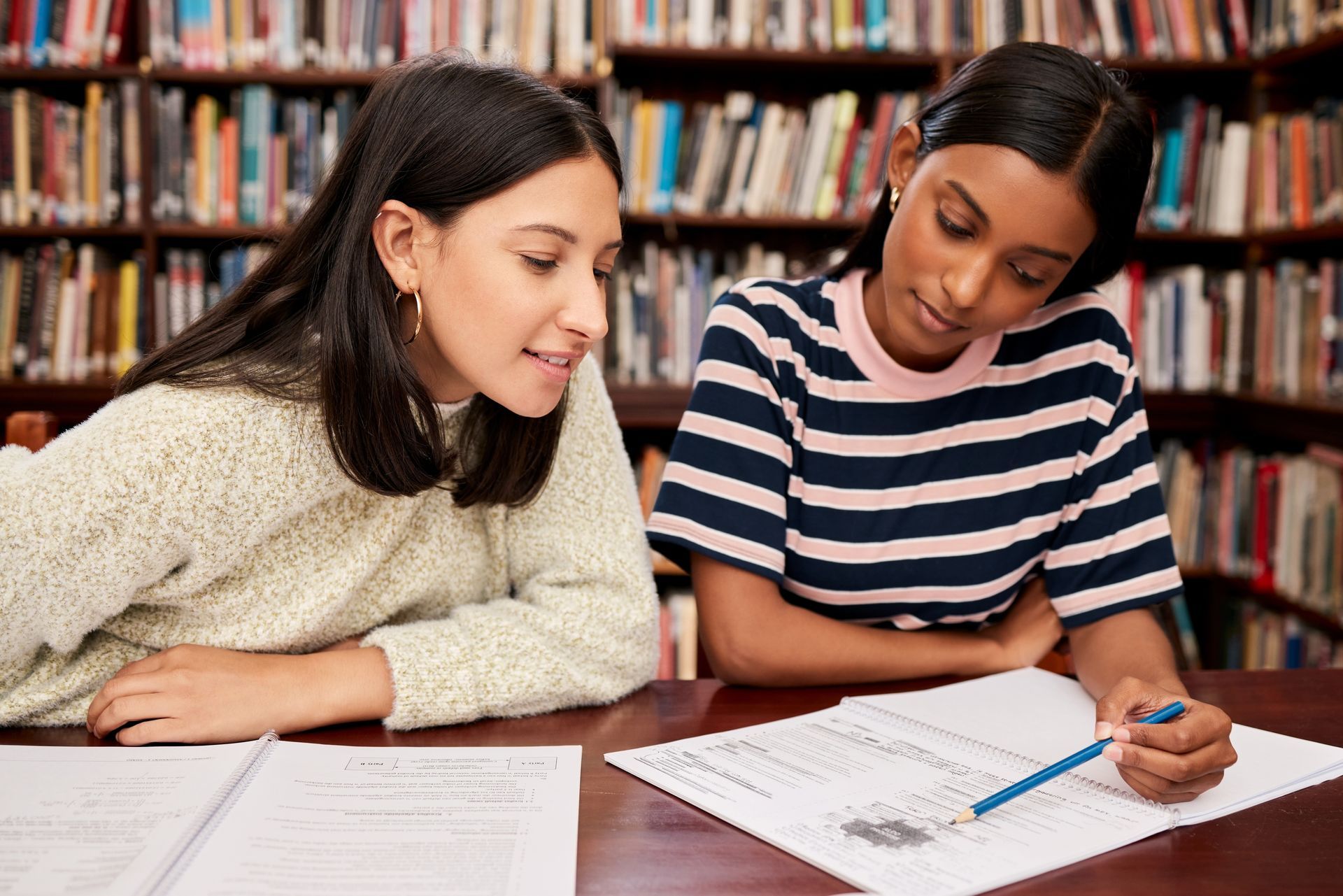Two young women are sitting at a table in a library looking at a piece of paper.