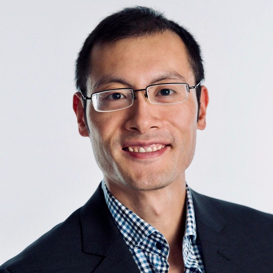 Chi Chen Man wearing glasses, smiling, with a checkered shirt and dark blazer against a white backdrop.
