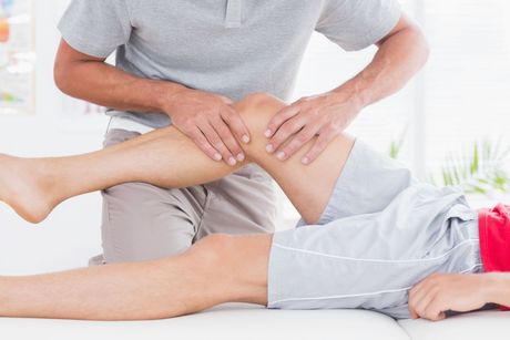 Physiotherapy Therapist examines patient's knee, applying pressure. Both are in a bright room.