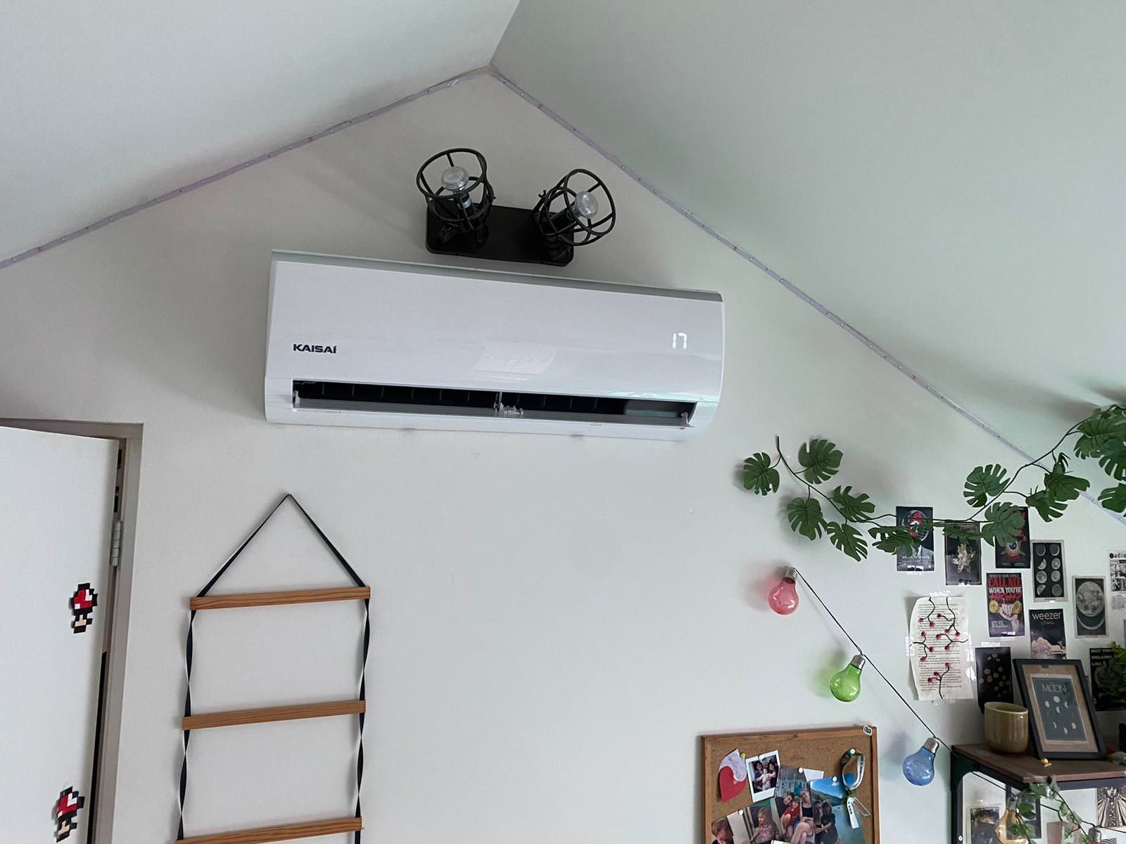 Witte airconditioner aan de muur met decoratieve elementen: klimplanten, foto's en een houten plank.