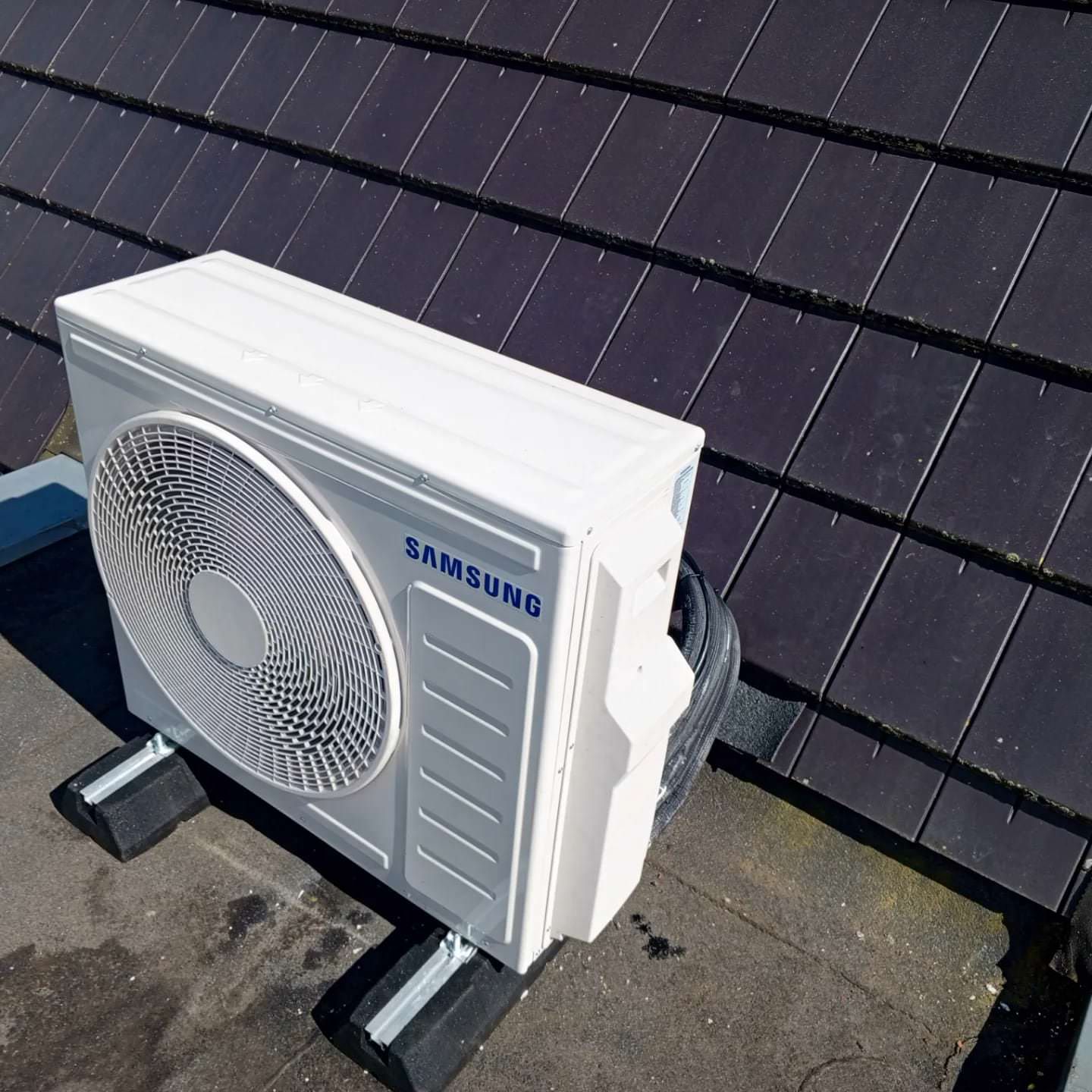 Een Samsung airconditioner staat op een donker pannendak, rustend op zwarte steunen.