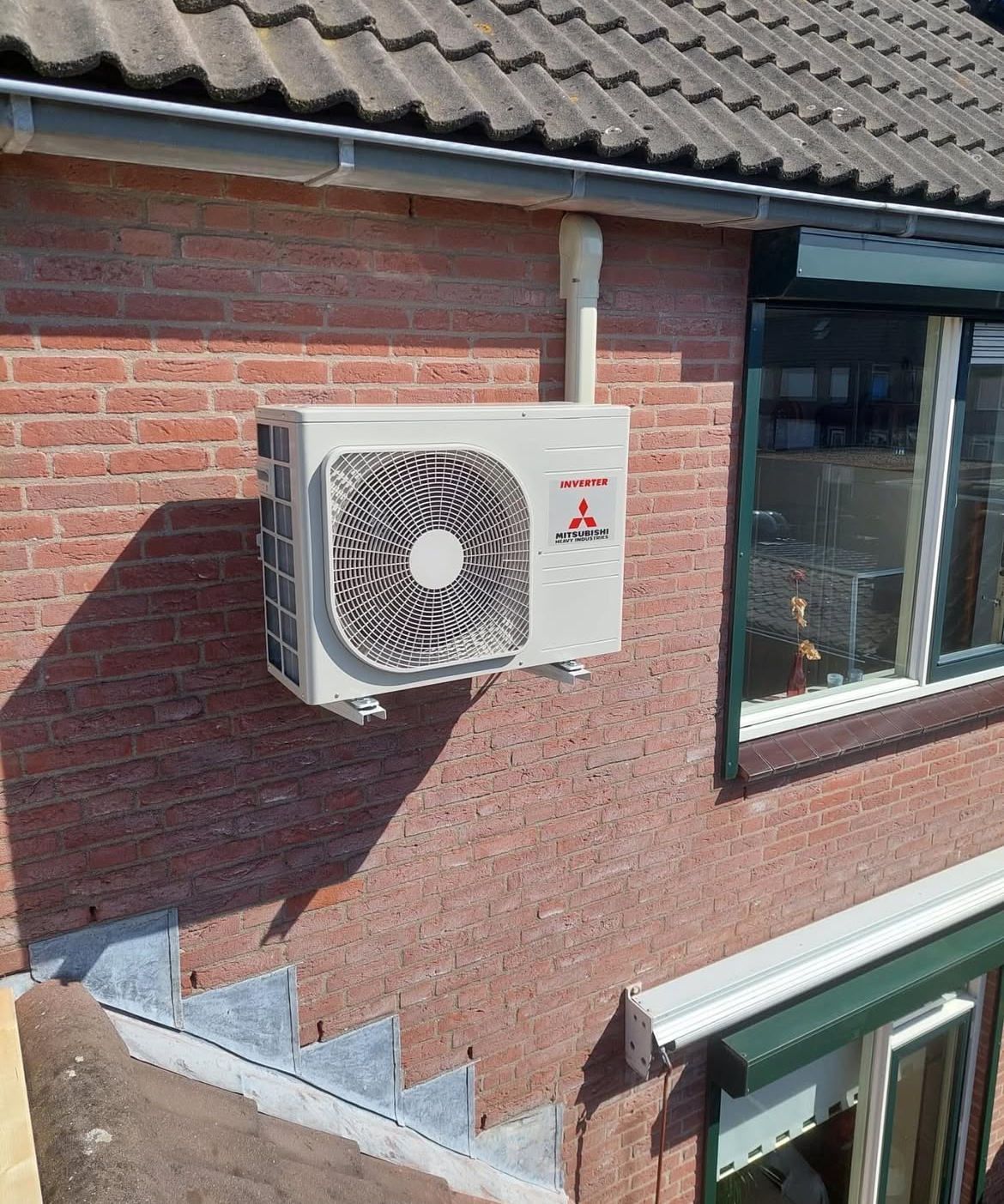 Mitsubishi airconditioningunit gemonteerd aan een rode bakstenen buitenmuur vlakbij een raam.