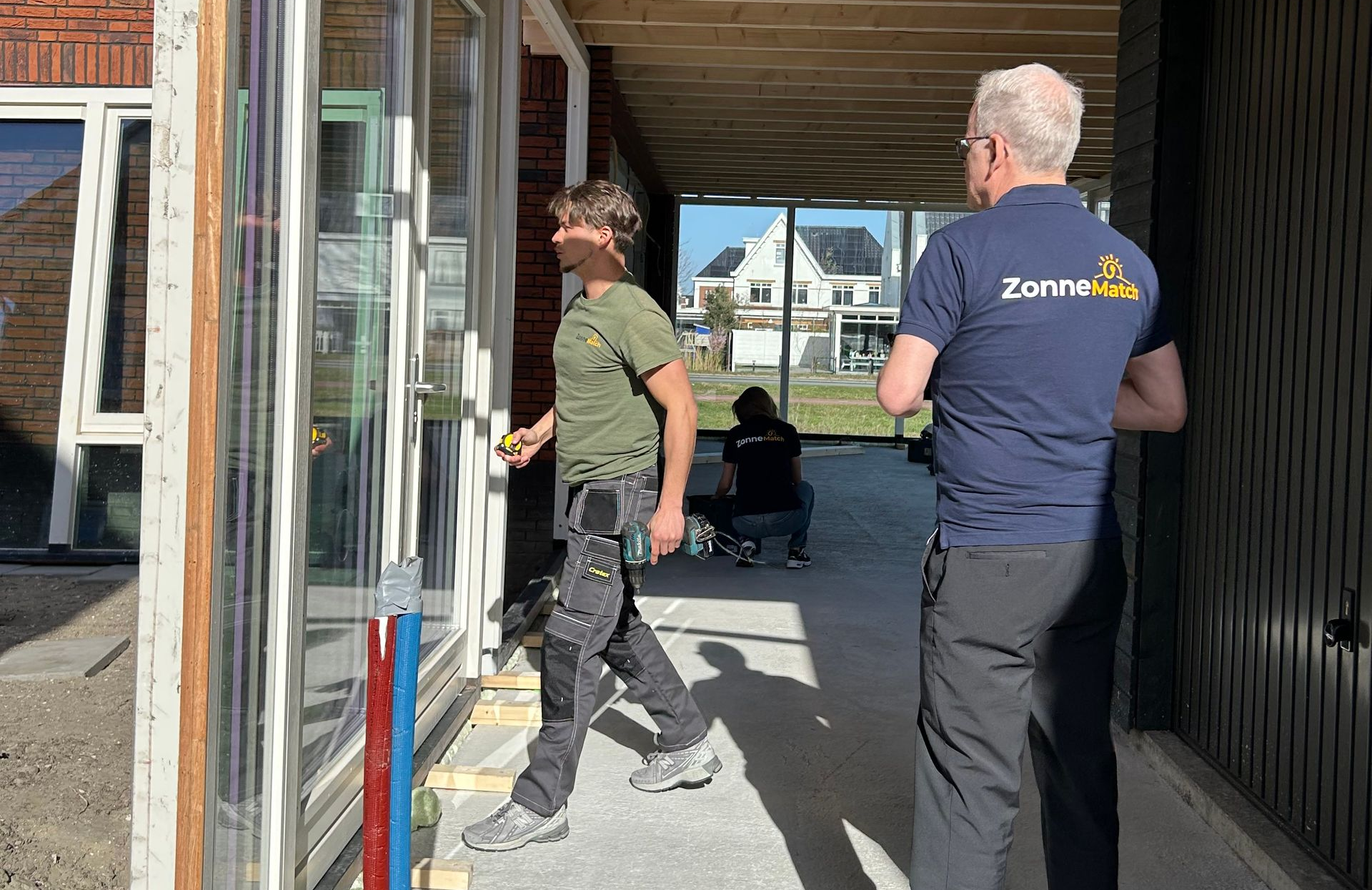 Werknemers bij een glazen deurkozijn: een opent de deur, een ander kijkt toe; in de entree van een gebouw.