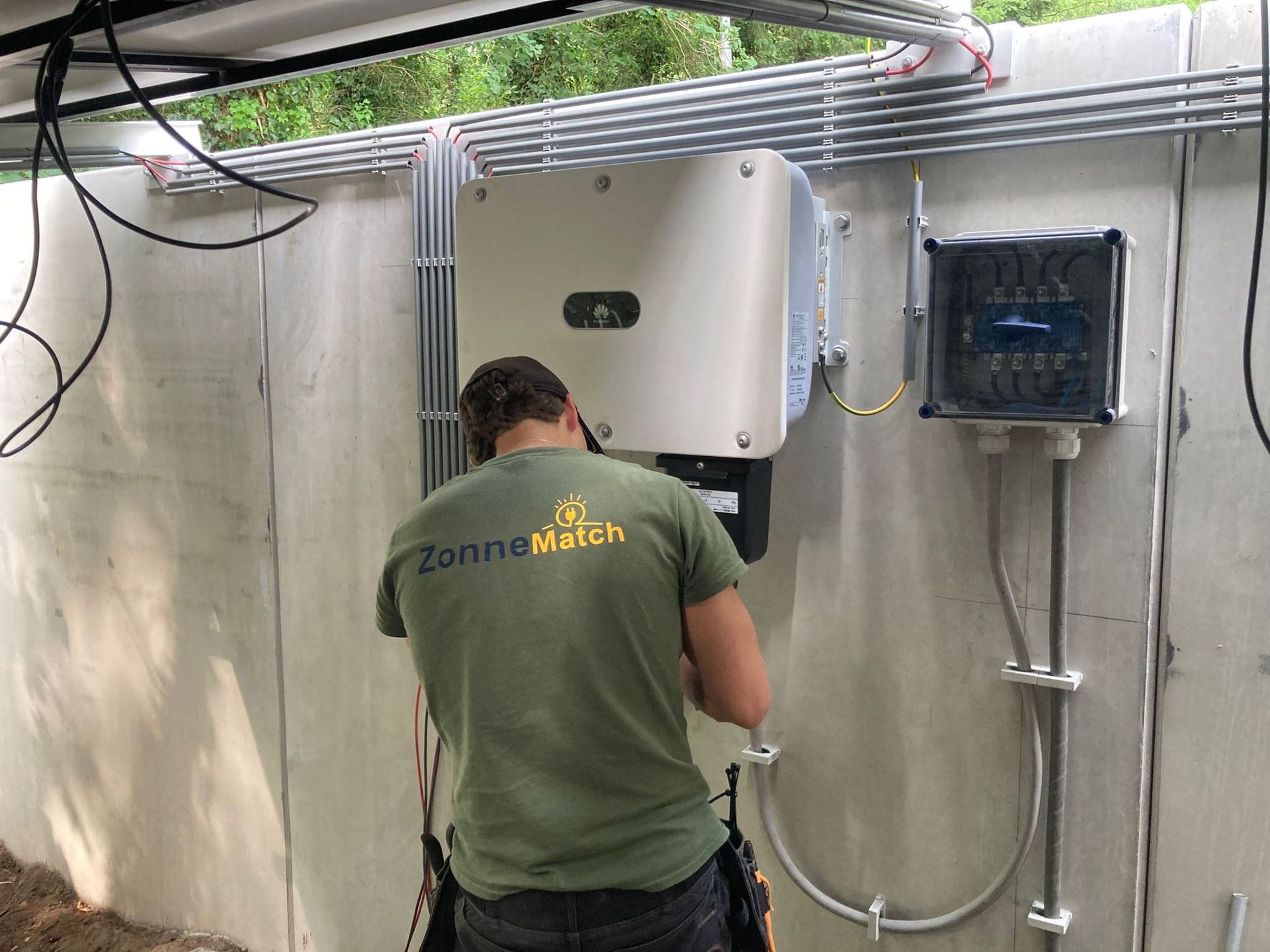 Persoon in groen shirt werkt aan zonnepanelen op een betonnen muur. Draden en elektrische verdeeldozen zijn zichtbaar.