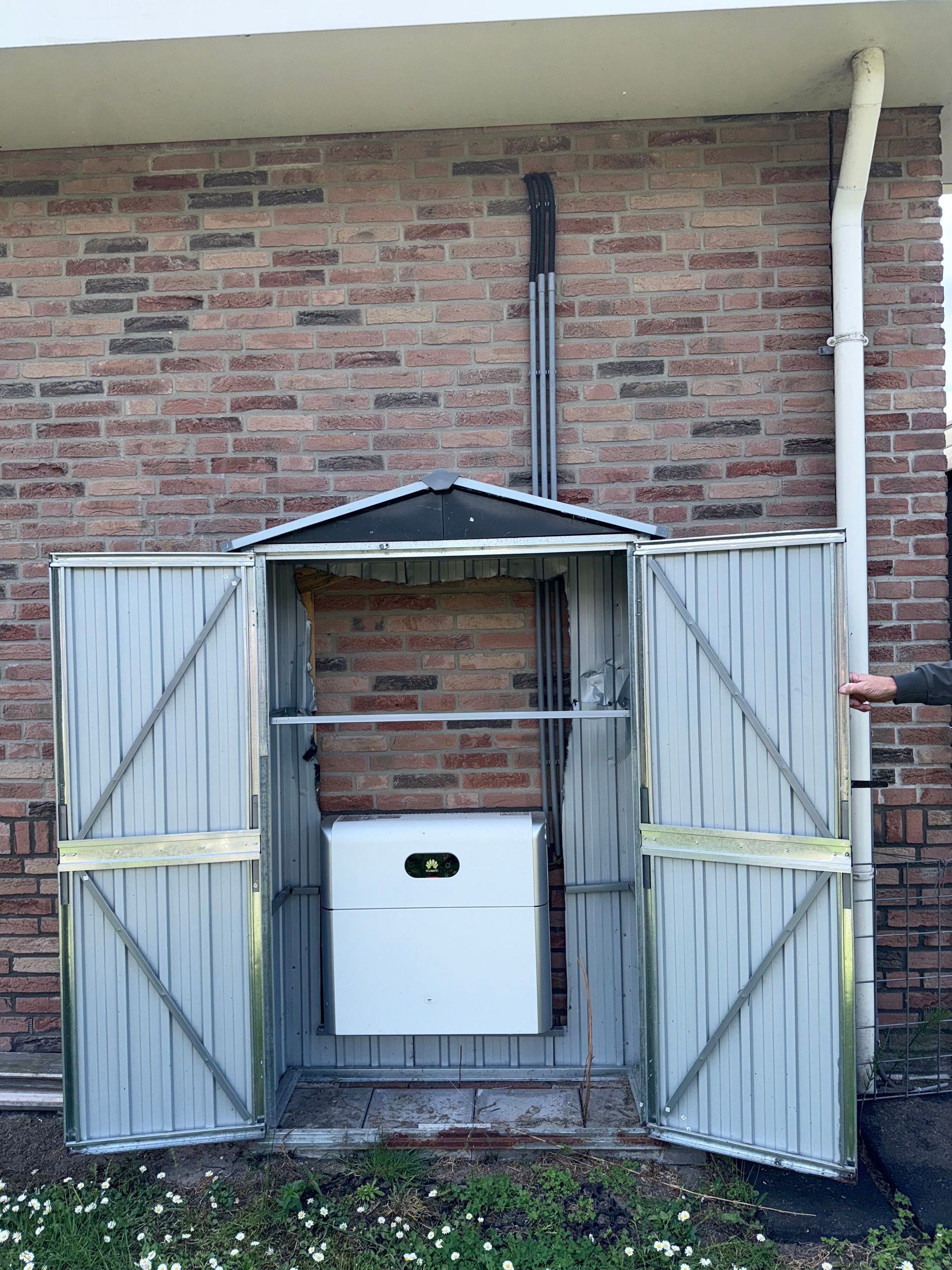 Metalen schuur met open deuren tegen een bakstenen muur, met daarin een witte doos.