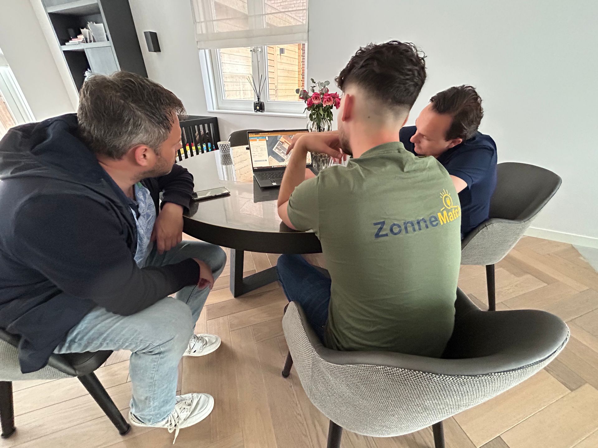 Drie mensen zitten aan een tafel, twee kijken naar een laptop, één draagt ​​een groen shirt met een logo, in een lichte ruimte.