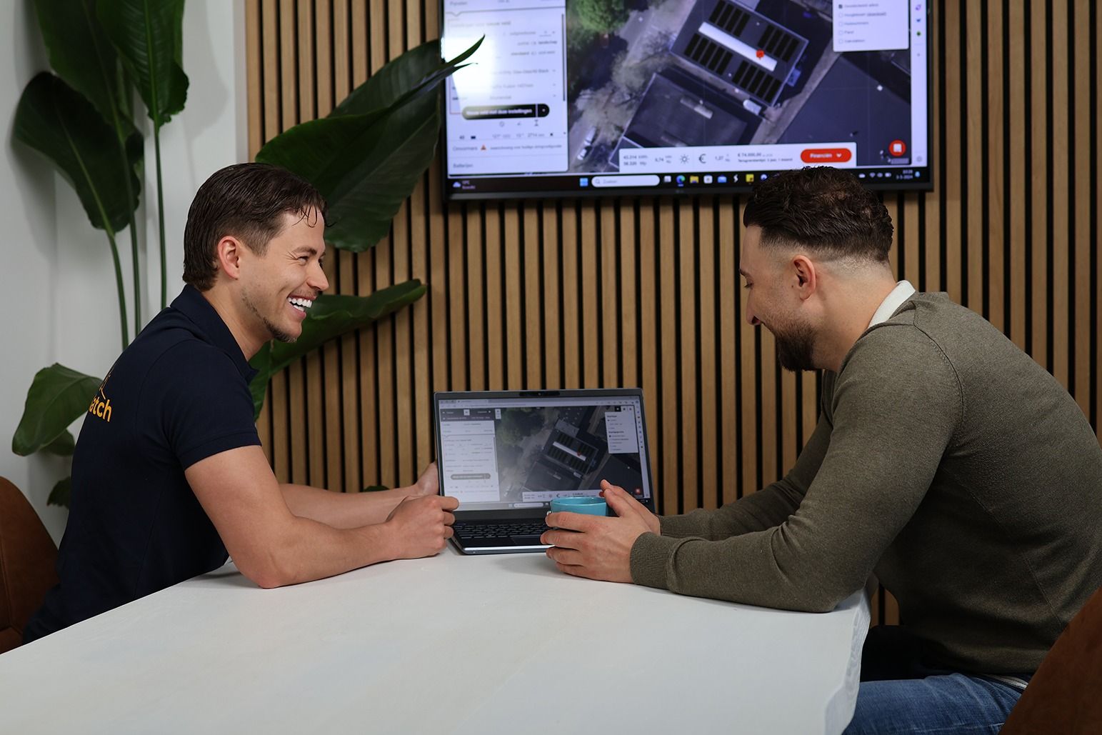 Twee mannen glimlachen en kijken naar een laptop, met een scherm waarop een kaart te zien is, een houten achtergrond en een plant.