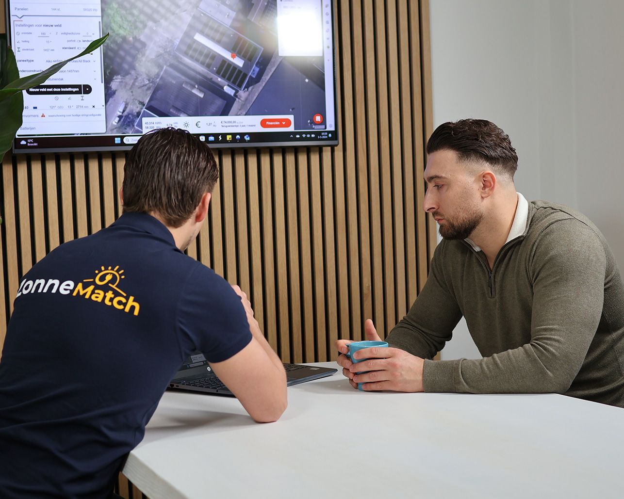 Twee mannen bespreken iets in een kantoor; op de achtergrond is een scherm te zien met een luchtfoto. De ene draagt een blauw shirt, de andere een groene trui.