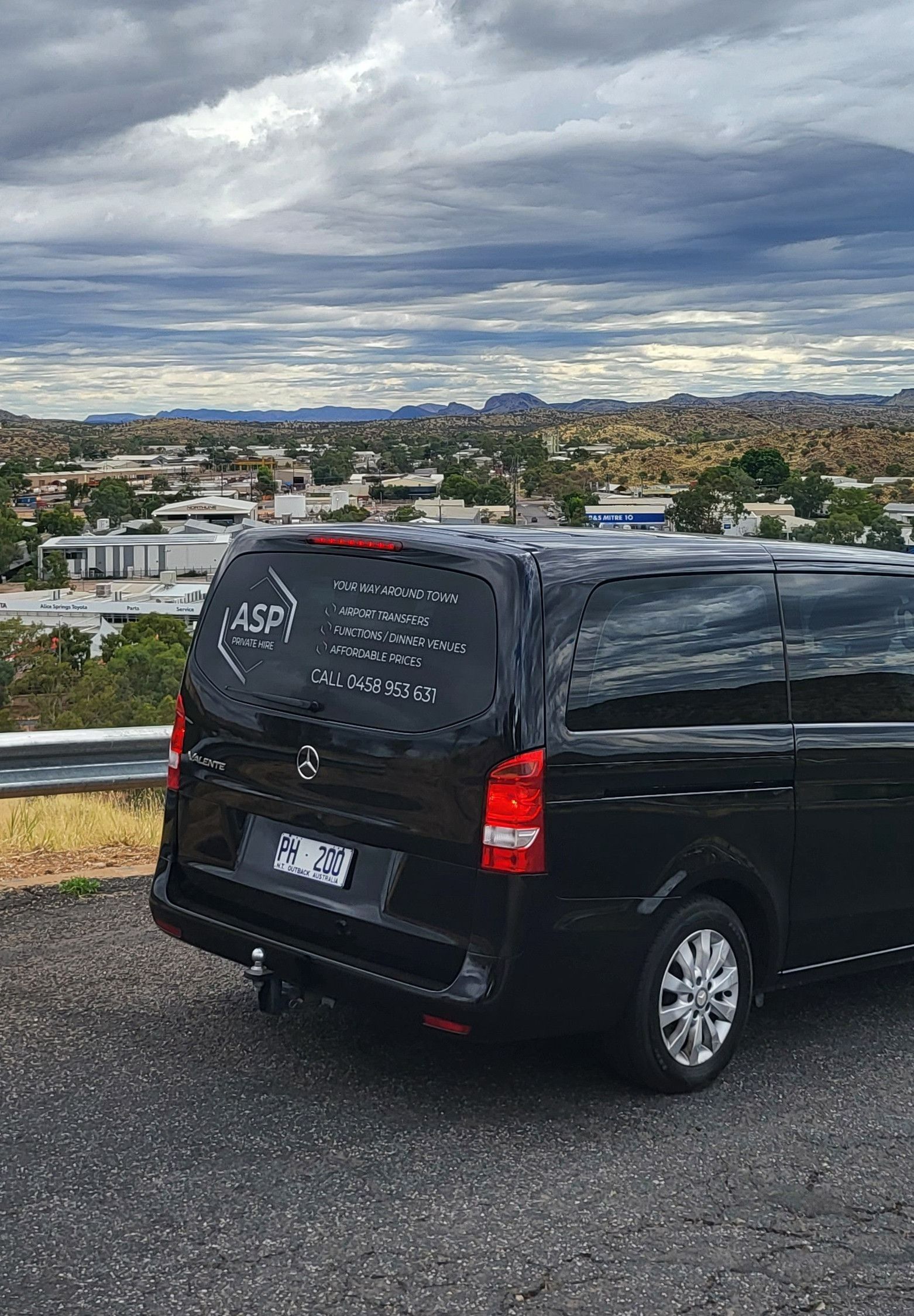 Black van parked on a gravel overlook with a cloudy cityscape and hills in the background — ASP Private Hire in Ross, NT