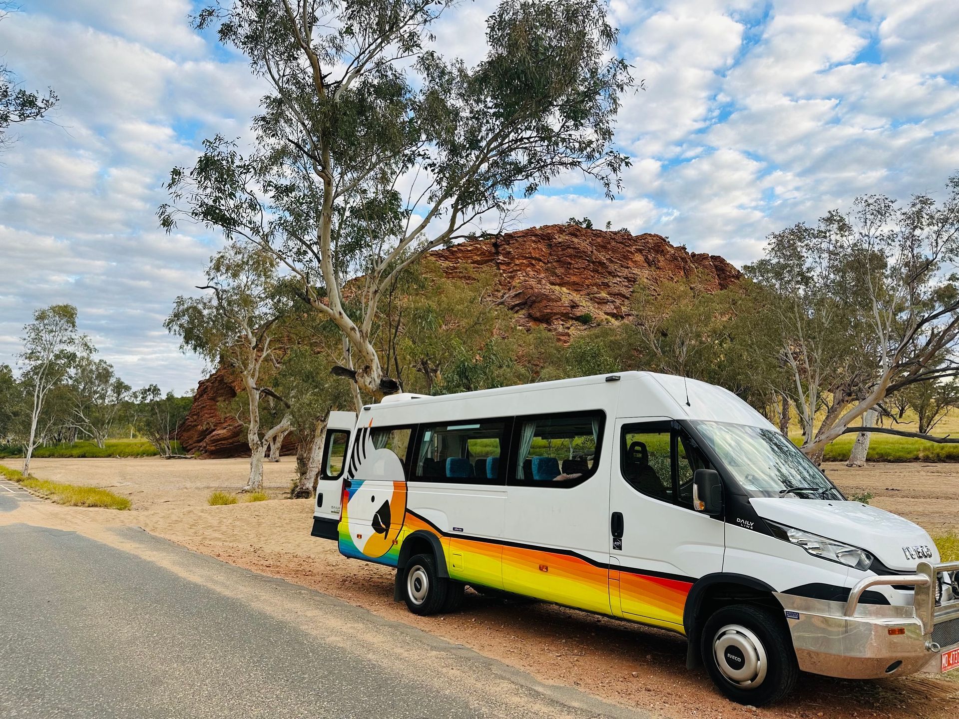 White shuttle bus parked on a roadside beside trees and a rocky hill under a cloudy sky — ASP Private Hire in Ross, NT
