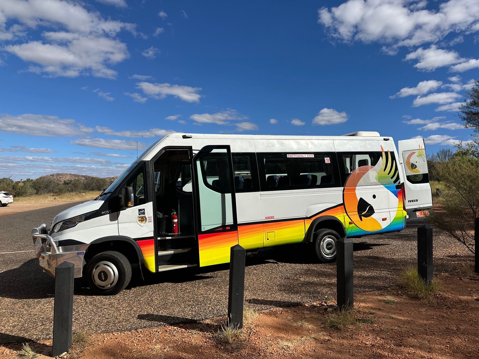 White minibus with colourful rear mural parked on gravel under a blue sky, beside black posts — ASP Private Hire in Ross, NT