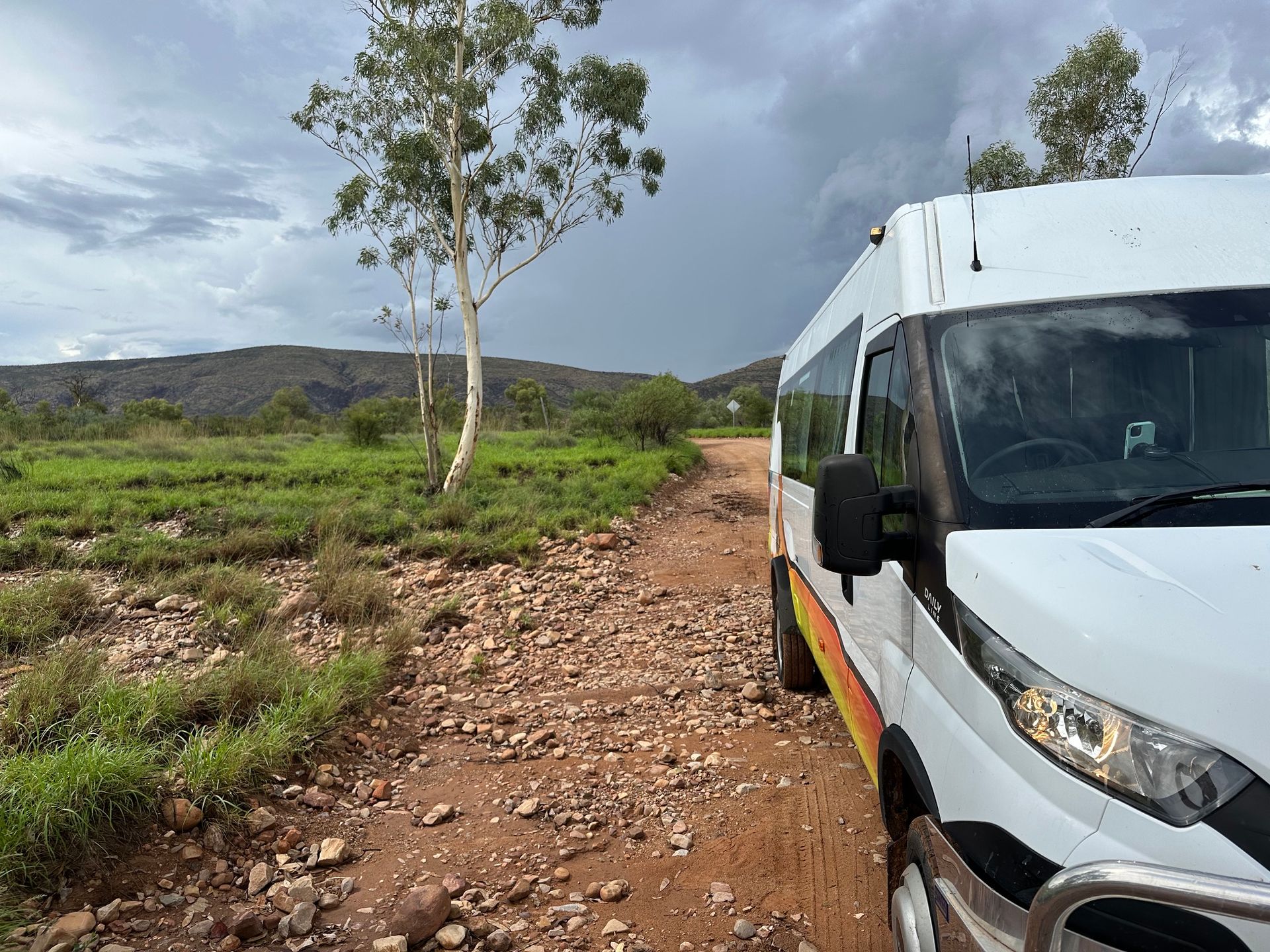 White van on rocky dirt road beside green field under cloudy sky — ASP Private Hire in Ross, NT