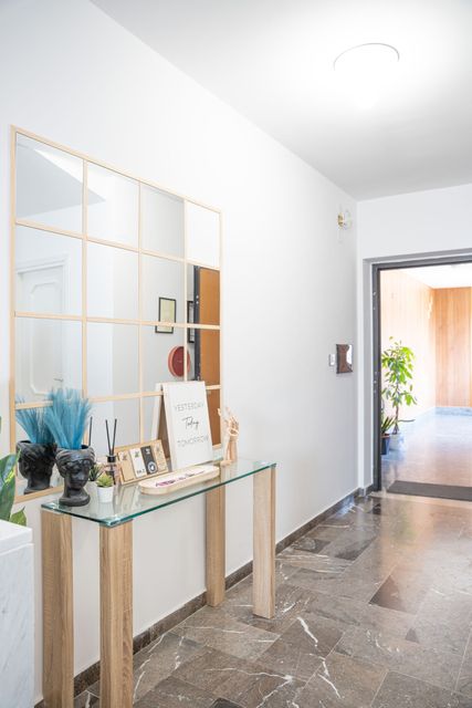 Corridoio moderno con consolle in vetro, specchio dorato e porta aperta su una luminosa area esterna.