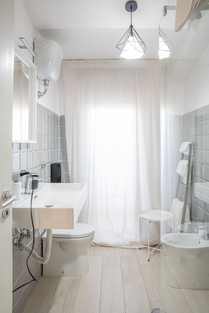 Bagno luminoso e bianco con lavabo, WC, doccia e tende trasparenti a una finestra.