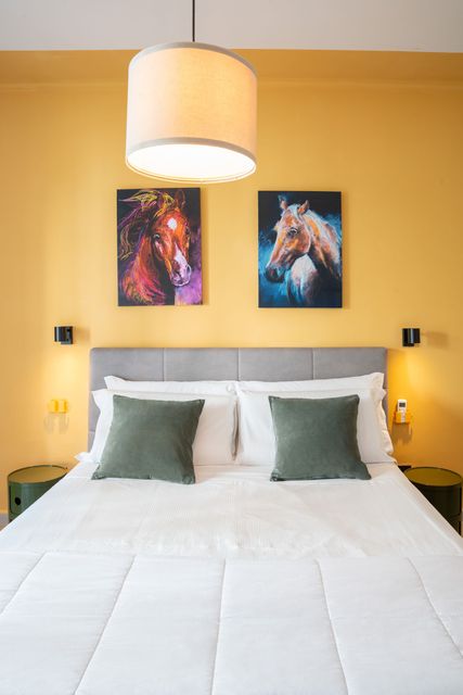 Camera da letto luminosa con letto bianco, cuscini verdi, pareti gialle e due quadri di cavalli sopra la testiera.