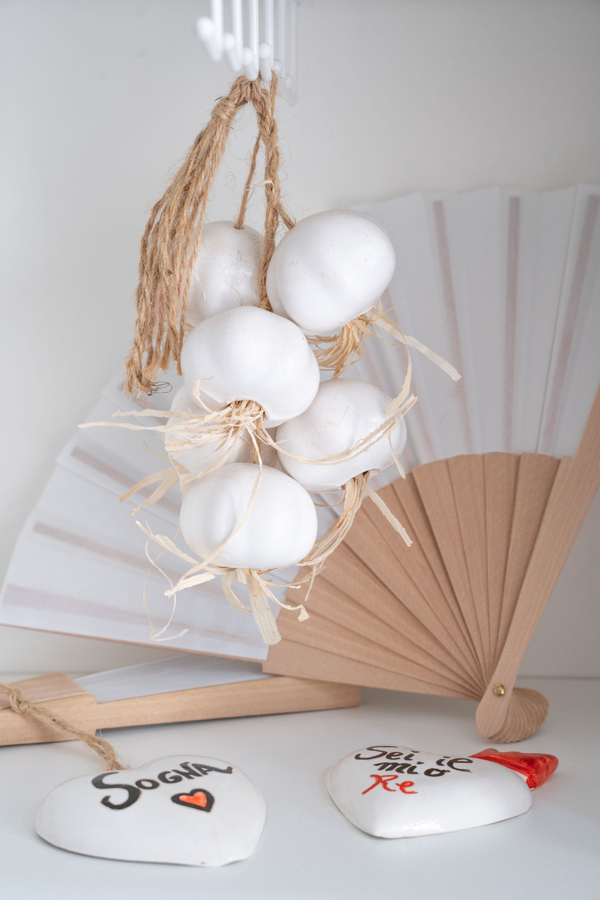 Ventagli decorativi bianchi e beige con ornamenti bianchi pendenti e cartoncino bianco su sfondo chiaro.