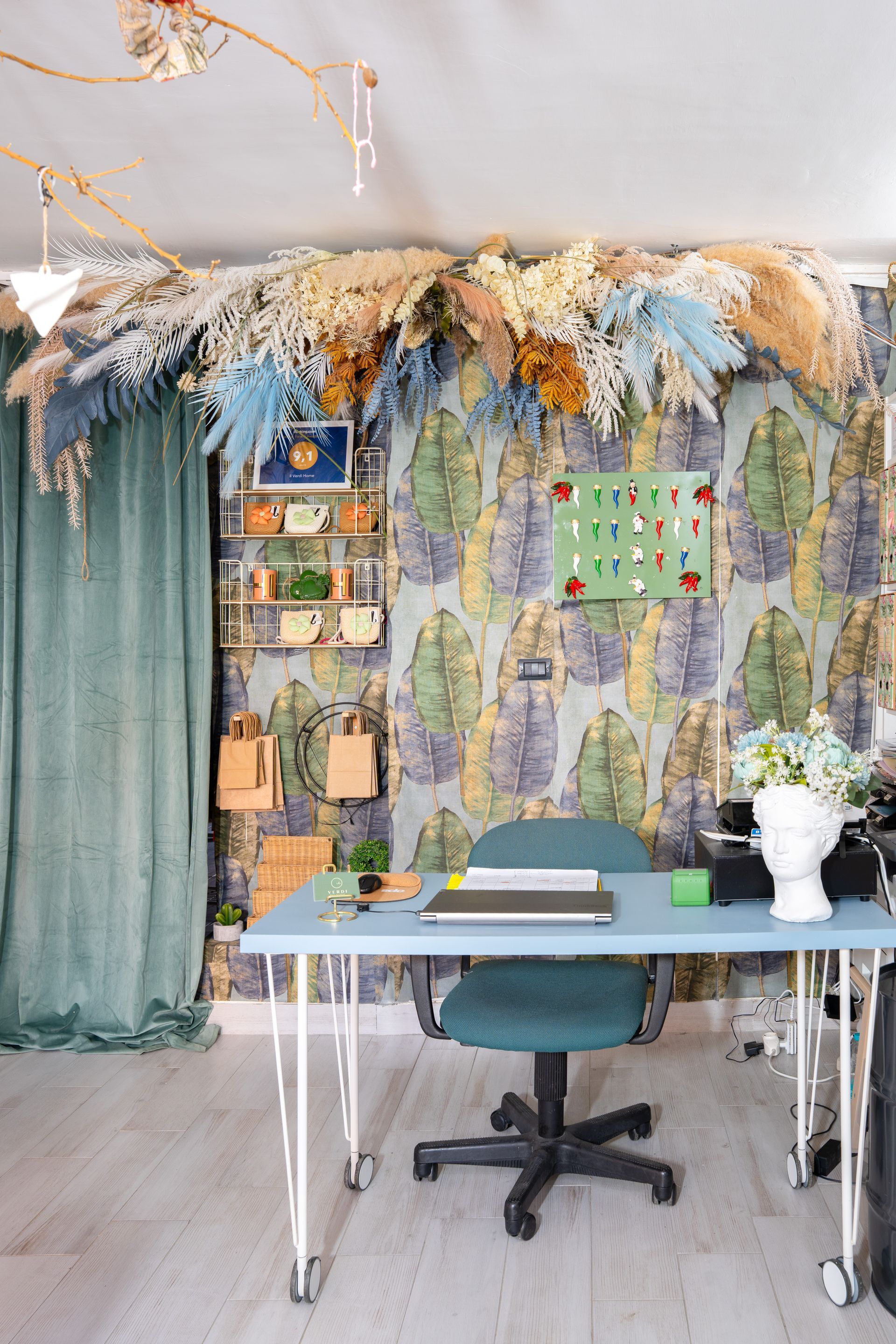 Ufficio in stile boho con scrivania, sedia color verde acqua, tende verdi, decorazioni floreali alle pareti e fiori secchi appesi.