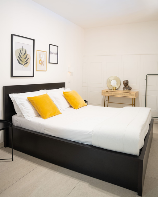 Camera da letto luminosa con letto nero, lenzuola bianche, cuscini gialli, quadri incorniciati e una piccola cassettiera.