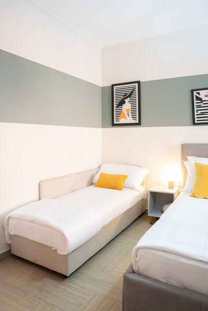 Luminosa camera doppia con due letti, lenzuola bianche, cuscini gialli e quadri alle pareti.