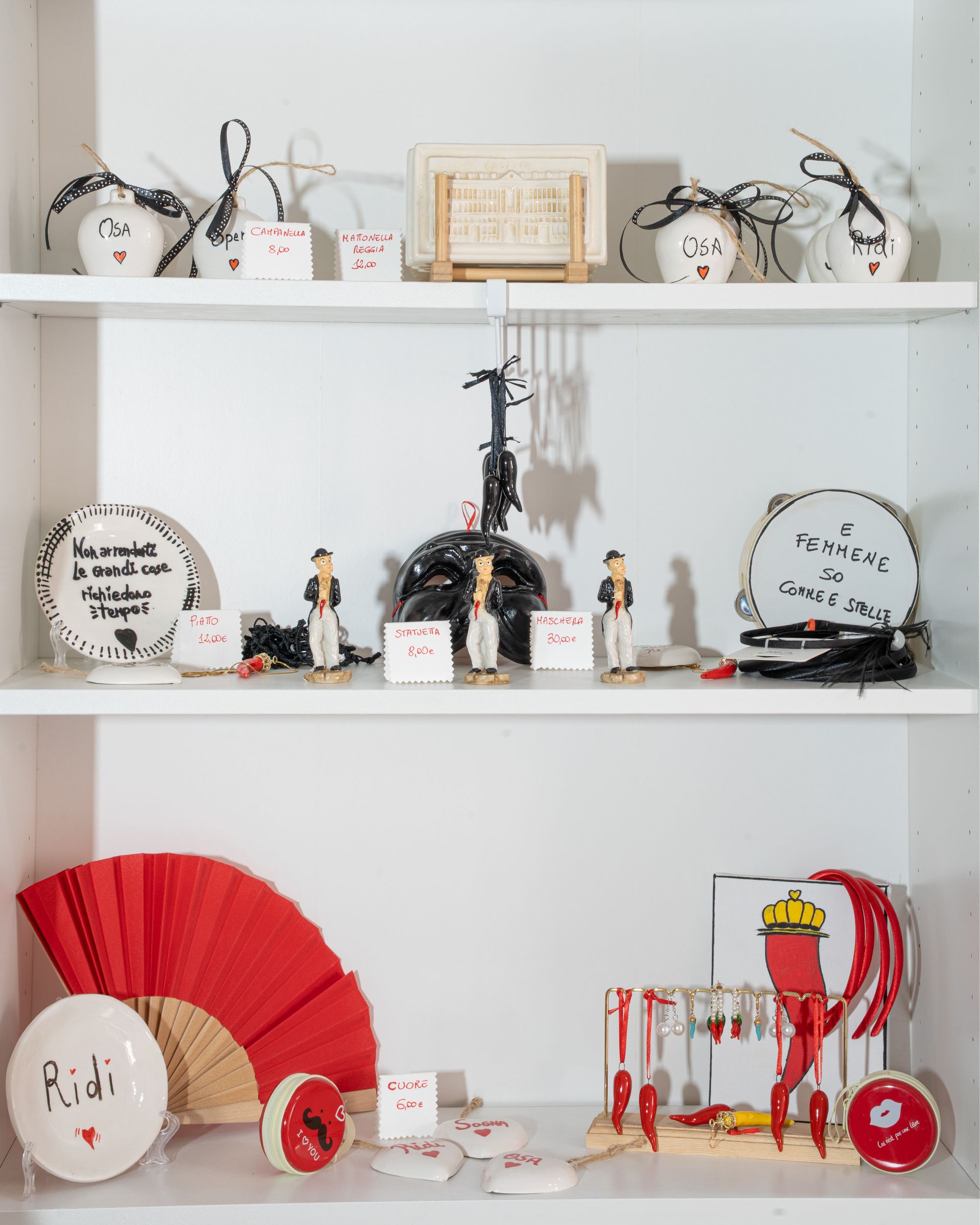 Su scaffali bianchi disposti su tre livelli, sono esposti piccoli oggetti decorativi, ornamenti e ventagli di carta rossa.