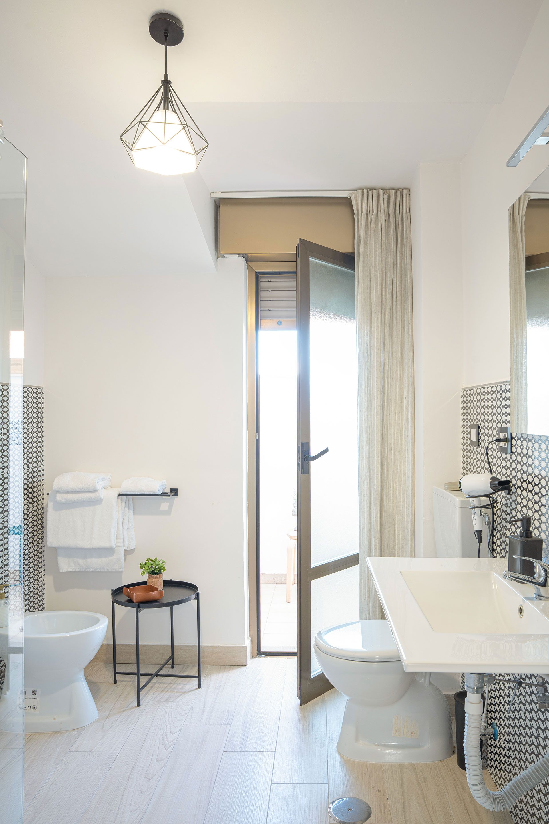 Luminoso bagno con WC, lavabo, doccia e pavimento in piastrelle bianche, visto dalla porta.