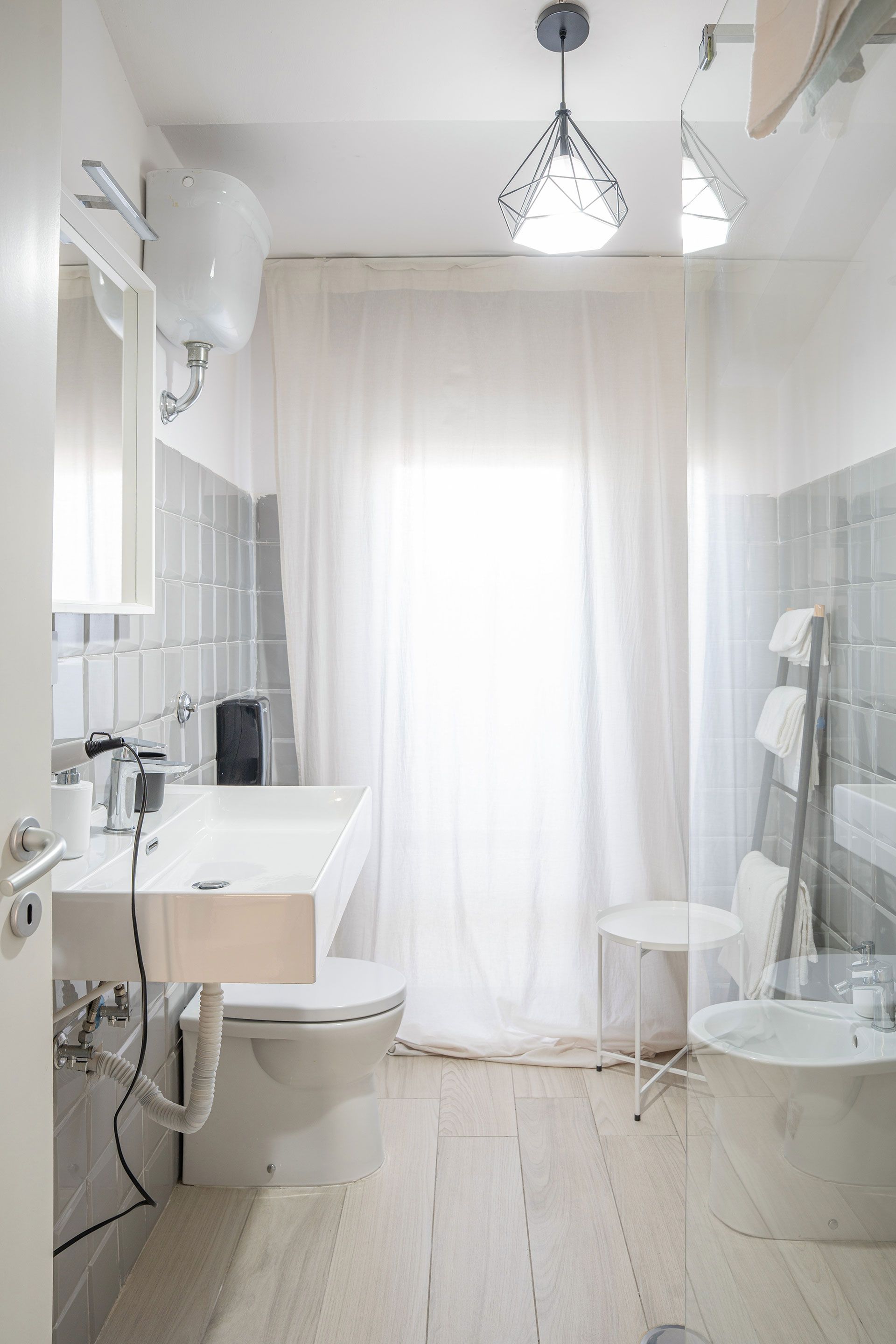 Bagno luminoso e bianco con vasca da bagno, lavabo, doccia e tende trasparenti a una finestra.